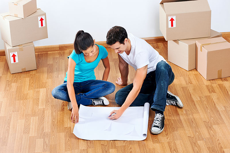 A couple sitting on the floor of an empty apartment with boxes around them looking at a floorplan